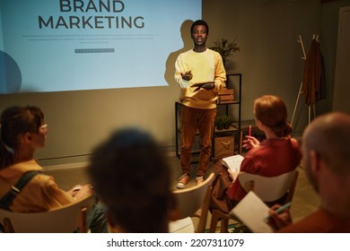 Portrait Of Young Black Man Giving Presentation On Brand Marketing To Team While Standing By Projector Screen And Addressing Audience In Business Training