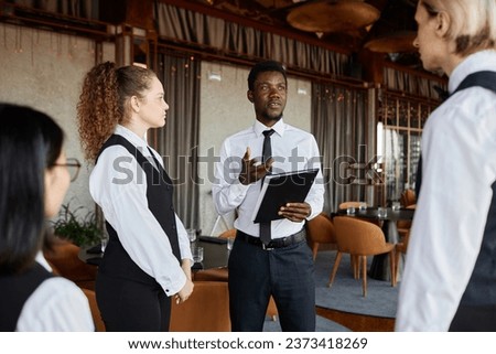 Portrait of young Black man talking to servers wearing classic uniforms during staff meeting in restaurant