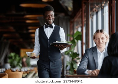 Portrait of young Black man as waiter bringing food on tray to guests enjoying dinner in luxury restaurant - Powered by Shutterstock