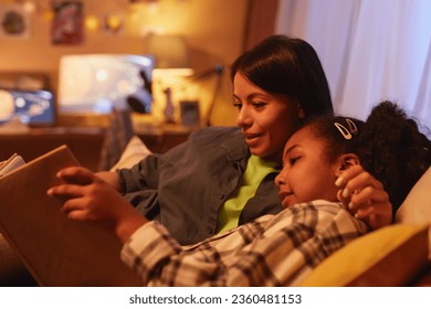 Portrait of young Black mother and daughter lying in bed together and reading book at night - Powered by Shutterstock