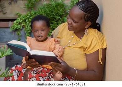 Portrait of a young black mother reading to her baby daughter sitting on her lap - Powered by Shutterstock