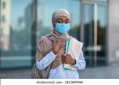 Portrait of young black muslim female student wearing medical mask posing outdoors with backpack and workbooks, resting at campus, following quarantine measures to prevent covid-19 outbreak - Powered by Shutterstock