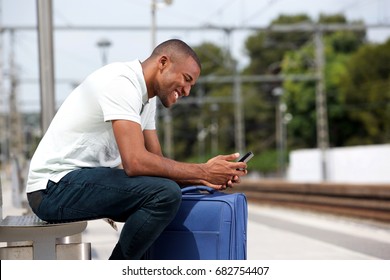 Portrait of young black traveler man with suitcase and using smart phone at railway platform