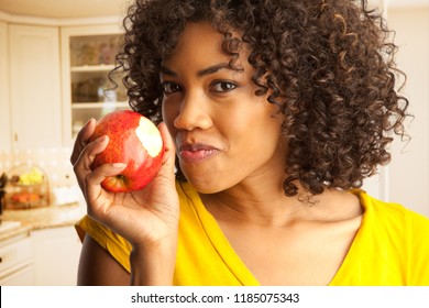 Portrait Of Young Black Woman Eating Fresh Red Apple Inside House Kitchen