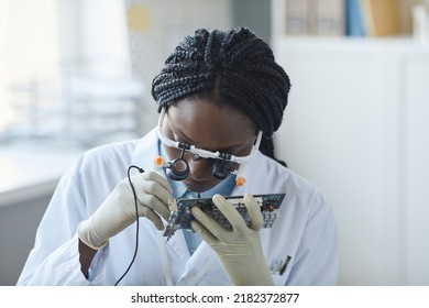 Portrait of young black woman inspecting computer parts and wearing magnifying visor while working in engineering lab - Powered by Shutterstock