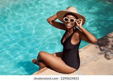 Portrait of young Black woman sitting with legs in pool, holding sunhat posing - Powered by Shutterstock