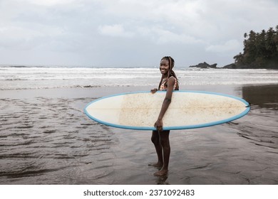 Portrait of a young black woman surfer holding her surfboard and smiling confidently standing on the beach in Sao Tome and Principe, Africa - Powered by Shutterstock