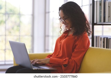 Portrait Of A Young Black Woman Wearing Orange Shirt Smiling And Using Laptop For Shopping Online, Leaning Online, And Video Call On Sofa At Home. Business Women Work From Home.