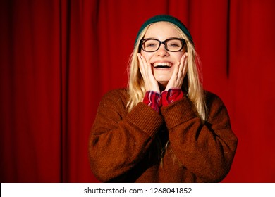 POrtrait Of Young Blond Hipster Woman In Eyeglasses Performs Stand Up Comedy And Joking At A Theater Over Red Backstage On Background