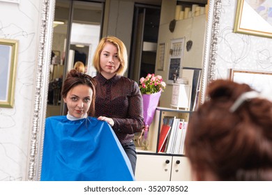 Portrait Of Young Blond Stylist Standing Behind Young Brunette Female Client Wearing Blue Smock And Sitting In Chair In Salon - Reflection In Large Mirror