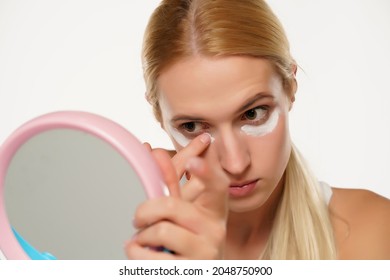 portrait of a young blond woman applying cosmetic product under her eyes on a white background - Shutterstock ID 2048750900