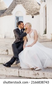 Portrait Of Young Blonde Bride In White Dress And Brunet Groom In Suit Looking At Each Other, Kissing And Sitting On Stairs Near Old City House In Italy. Beautiful And Romantic Wedding, Happy Couple