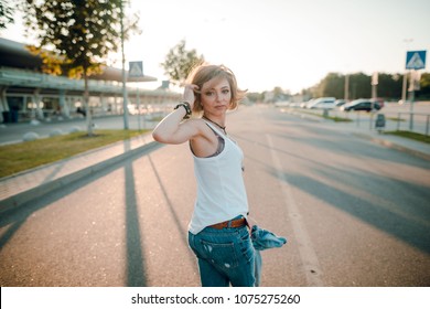 Portrait of a young blonde caucasian woman in blue jeans and white shirt walking on the street and turning her head. Evening sunset. Rear view