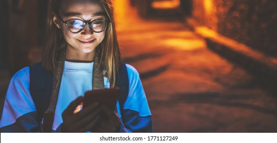 Portrait Young Blonde Girl With Reflection Of Glare In Glasses From Cellphone Screen Looking At Mobile Gadget At Night On Street Of Old Historical City Background Of Evening Lights, Blank Copy Space