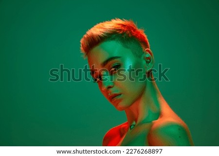 Portrait of young blonde girl with short hair and bare shoulders posing against green studio background in orange neon light. Cyberpunk culture. Concept of technology, modern fashion, futurism