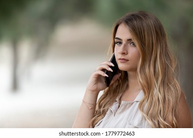 Portrait of young blonde italian woman talking at the phone inside the park whit shallow depth of field with blurred tree in the background