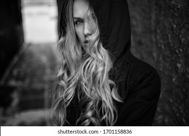 Portrait of a young blonde with long curly hair against a dark background. A mysterious girl with a hood on her head looking around the camera. Black-and-white portrait