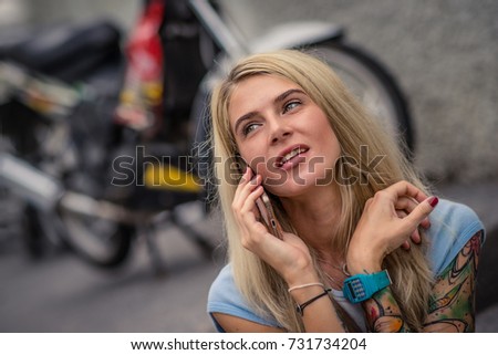 Portrait of young blonde sitting on the background of the motorcycle. Talking on the phone. Female hands with a tattoo. Urban fashion.