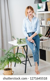 Portrait of a young blonde smiling attractive designer at cozy small studio. Tailor dressed modestly for work, wearing jeans and blue shirt. She has bare feet and she leaning her back against a table.