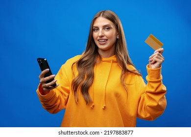 Portrait Of A Young Blonde Woman Showing Plastic Credit Card While Holding Mobile Phone Over Blue Background