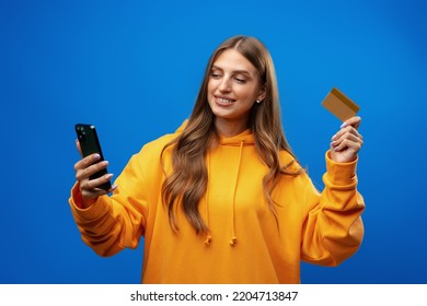 Portrait Of A Young Blonde Woman Showing Plastic Credit Card While Holding Mobile Phone Over Blue Background