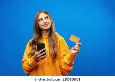 Portrait Of A Young Blonde Woman Showing Plastic Credit Card While Holding Mobile Phone Over Blue Background