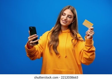 Portrait Of A Young Blonde Woman Showing Plastic Credit Card While Holding Mobile Phone Over Blue Background