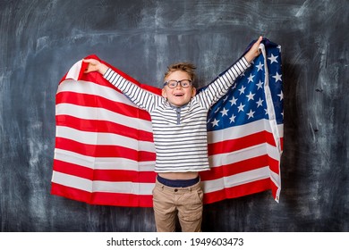 Portrait Of A Young Boy With An American Flag On Black Background. Kid In Glasses With Raised Hands Against Blackboard.