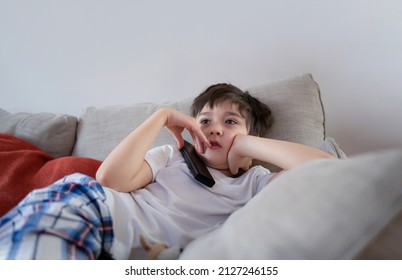 Portrait Young Boy With Curious Face Sitting On Sofa Watching Cartoon, School Kid Sitting Alone On Couch Holding Remote Control Watching TV, Child Relaxing In Living Room After Back From School.