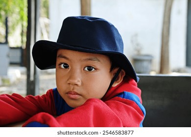 Portrait of a young boy wearing a dark blue hat and red hoodie, looking directly at the camera with a calm expression. The soft background highlights the boy's thoughtful gaze and natural innocence. - Powered by Shutterstock