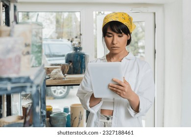 Portrait of young brunette asian female artisan in headscarf and workwear using digital tablet while standing in blurred ceramic workshop at background, creative process of pottery making - Powered by Shutterstock