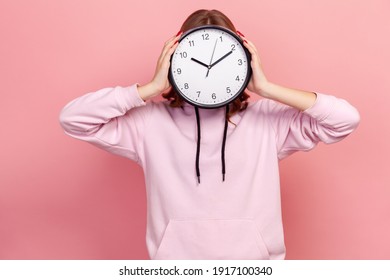 Portrait Of Young Brunette Female In Hoodie Holding Wall Clock Hiding Her Face, Time Management, Schedule And Meeting Appointment. Indoor Studio Shot Isolated On Pink Background