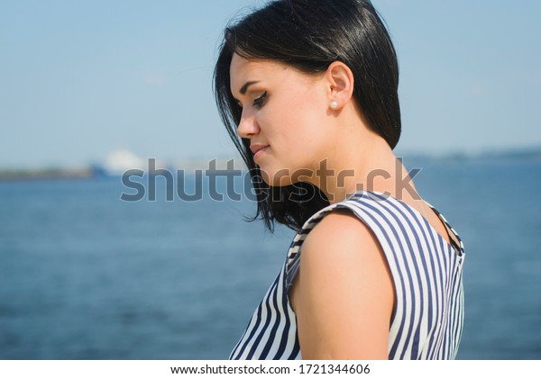 portrait of a young brunette\
girl of European descent, in a striped dress on a background of sea\
or river
