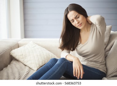 Portrait of a young brunette girl sitting on the couch at home with a headache and back pain.