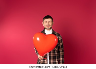 Portrait Of A Young Brunette Guy Looking To Camera, Holding A Heart-shaped Balloon On A Red Background. Holiday Concept. High Quality Photo