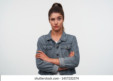 Portrait of young brunette lady with raised eyebrow wears in white t-shirt and denim jackets, looks at the camera with unhappy expression, stands over white background with crossed arms.