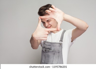 Portrait Of Young Brunette Man In Casual Style With White T-shirt And Denim Overalls Standing And Looking At Camera And Making Camera Frame With Hands. Indoor Studio Shot, Isolated On Gray Background.