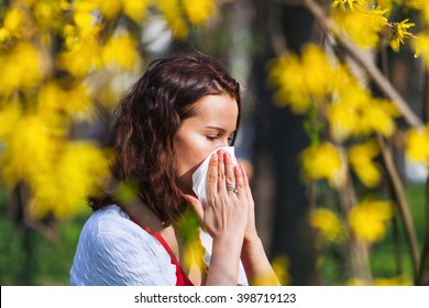 Portrait of a young brunette woman blowing her nose when standing close to flowers in a park
