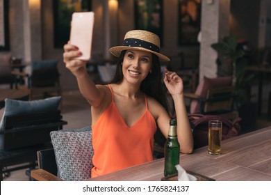 Portrait Of Young Brunette Woman Taking A Selfie Photo From Cell Phone In Pub Bar. 20s Hispanic Girl In Summer Hat And Clothes Using Mobile Phone To Video Call People In Indoors.