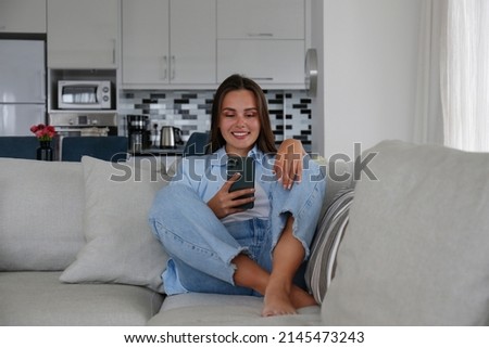 Portrait of young brunette woman wearing mom jeans lying on the couch texting and smiling. Joyful female having fun video phone call. Background, copy space, close up.