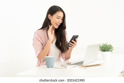 Portrait of young business Asian woman uses mobile phone for vdo call via 5G internet connection smile wave hand hello. During break from working in modern office. Mobile business technology concept