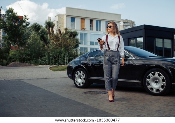 Portrait of young\
business boss woman standing near expensive luxury car with clutch\
bag, dressed in white shirt, trousers, suspenders, looking away.\
Image with copy space.