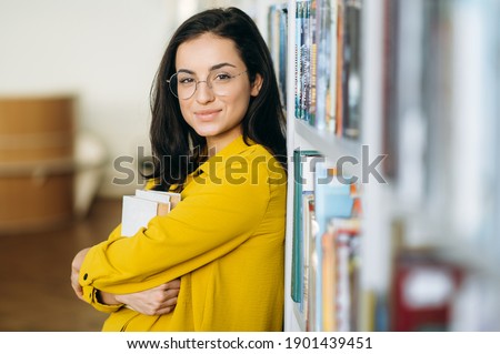 Portrait of young business female or student in eyeglasses, holding in arms some books, standing in library. Stylish caucasian female teacher looking at the camera, smiling, education concept