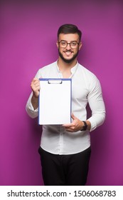 Portrait Of A Young, Business Man Standing Against A Plain Violet Background And Smiling While He Holds His Leather Folio. He Is Well-dressed In Business Casual And Looks Relaxed.