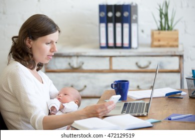 Portrait Of Young Business Mom Holding Her Newborn Cute Babe While Working In Home Office Interior, Looking At Cellphone Screen. Serious Working Mother Using Mobile Phone And Nursing New Born Child