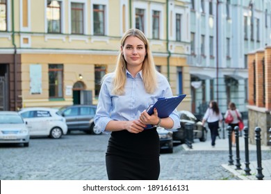 Portrait of young business woman with business papers, file folder, female looking at camera outdoors, city street background