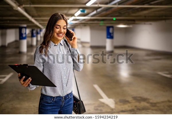 Portrait of young business woman with smart phone
in the underground car parking of the new residential building.
Businesswoman in underground garage. Elegant woman using smartphone
in parking garage