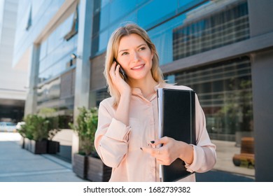 Portrait Of Young Business Woman Talking On The Phone While Standing Outside Office Buildings. Business And Success Concept.