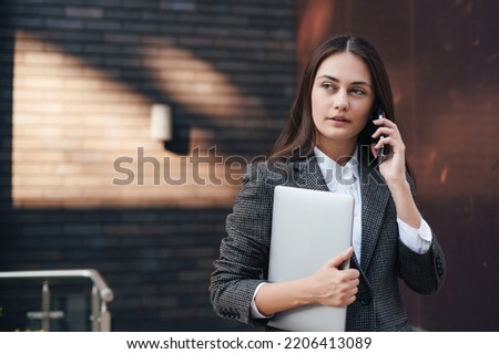 Portrait of young business woman talking on the phone, holding laptop while standing outdoors at the street on sunny day, joyful, relaxing. Making phone call