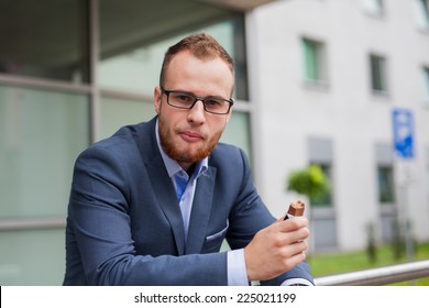 Portrait Of Young Businessman With Beard Standing In Front Of Office Block. He Is Eating Candy Bar. Outdoor Photo.